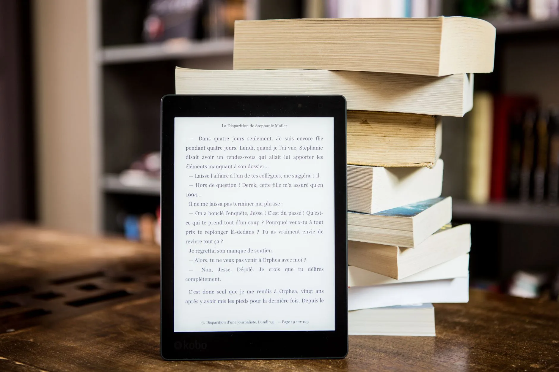 Isca digital ou ebook — Leitor de e-books com texto exibido ao lado de uma pilha de livros físicos em uma mesa de madeir